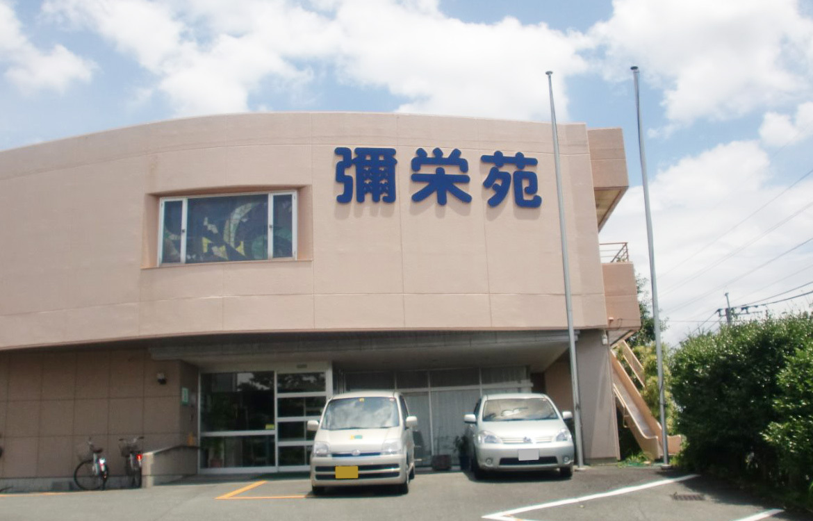 介護施設の建物外観。ベージュ色の外壁に施設名の看板が付いており、駐車場には車が停まっている。
