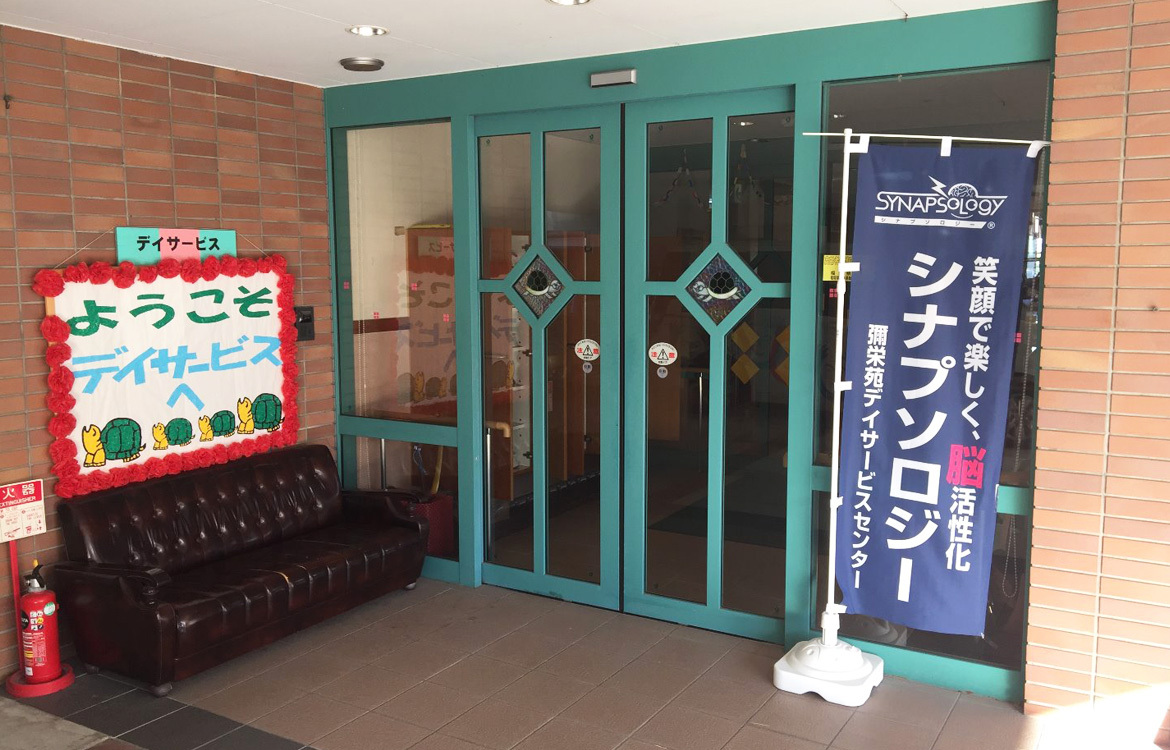 介護施設の玄関・受付エリア。デイサービスの案内看板と緑色のドアが見える。